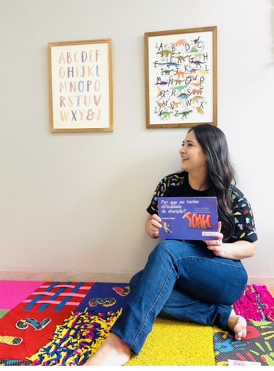 Daniella no consultório segurando o livro infantil sobre TDAH, com tapete sensorial colorido ao redor